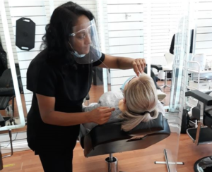 Mangala is wearing a PPE plastic face shield whilst threading the eyebrows of a client who is reclining in a chair below her.