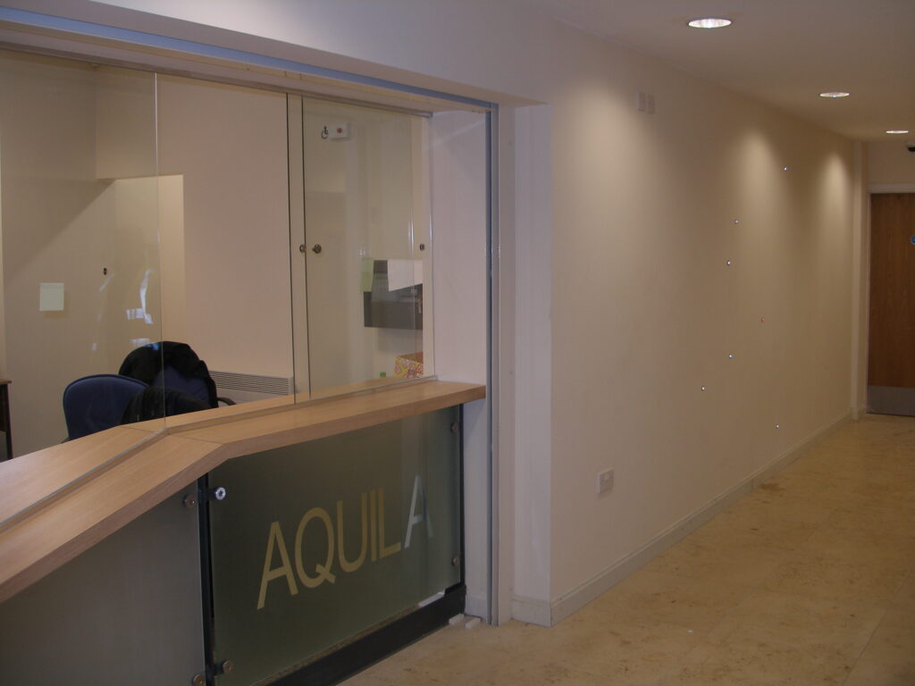 A reception area, glassed off and separate from the foyer. The foyer is tiled with pale, sandy coloured tiles. The label on the reception desk says "Aquila"