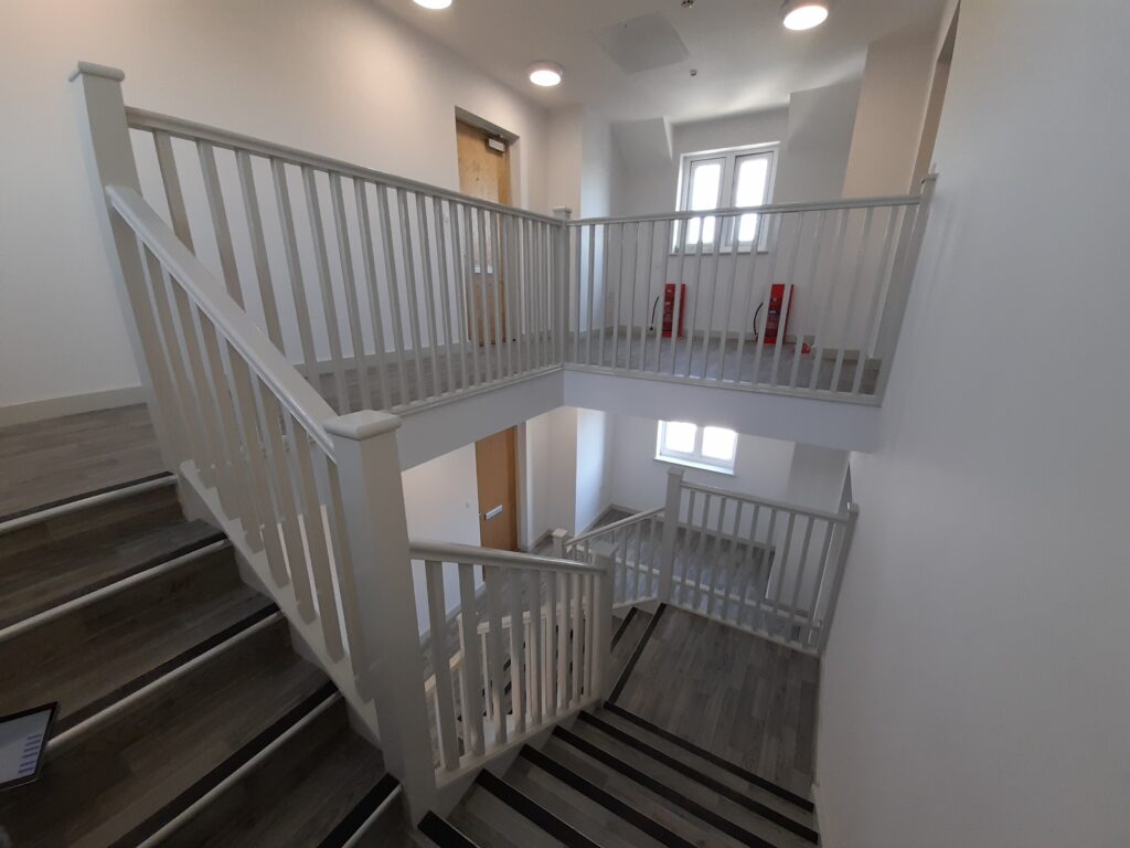 A stairwell with white banisters and grey flooring in a wooden effect. The vantage point displays two floors.