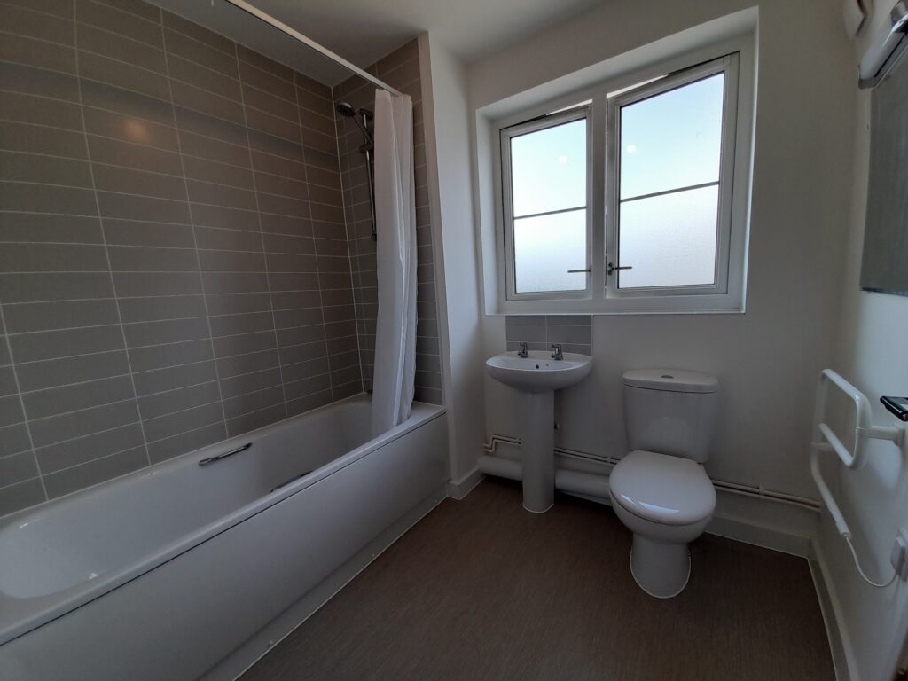 A bathtub, toilet, and sink are situated below a window. The bathroom has grey tiles and the floor is laminate.